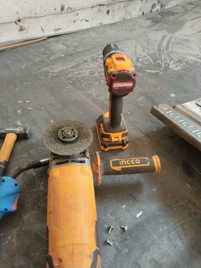The image shows a collection of tools on a concrete floor, likely in an outdoor or workshop setting. In the foreground, an orange angle grinder with a circular cutting disc is visible. To the left of the grinder, a hammer with an orange handle is laid out. A blue power tool, possibly a drill or sander, is partially visible to the left of the grinder. In the background, an orange and black cordless drill with "BRUSHLESS" written on it stands upright. To the right of the drill, a metallic object, possibly a piece of equipment or material, is present. The floor is dark and appears worn, with some scattered debris. The wall in the background is light-colored and shows signs of wear and possibly water damage. There are no people in the image. The scene depicts tools commonly used for construction or renovation work. The location is specified as Chalysh, Uzbekistan, which implies a setting within that region.