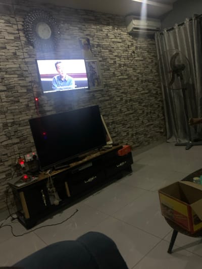 A television mounted on a textured gray wall displays a man in a blue shirt. Above the TV, framed pictures adorn the wall. A decorative mirror sits near the lower left corner. To the right of the TV, a long, black entertainment center holds various objects. Above the entertainment center, a cardboard box sits on a table. In the upper left, a ceiling fan is partially visible, along with an air conditioning unit. The floor is tiled.