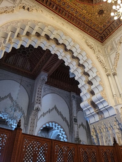 The image presents a detailed interior view of a grand structure in Bouznika, Morocco, consistent with traditional Moroccan Islamic architecture, likely a mosque or royal reception hall. The scene contains no human subjects or visible actions.

In the lower foreground, a dark brown wooden screen or balustrade with intricate geometric latticework forms a prominent partition. Above this, a large, multi-lobed or horseshoe arch constructed from a light-colored material, possibly carved plaster or stucco, dominates the midground. The underside of this arch features elaborate muqarnas (stalactite vaulting) in a white or light grey tone, creating a three-dimensional effect. The arch spandrels and surrounding wall surfaces are extensively decorated with fine, low-relief stucco carvings displaying intricate geometric and curvilinear arabesque patterns.

Further into the background, multiple additional arches are visible, receding into the space, each exhibiting similar decorative motifs on their surfaces. The high ceiling is richly ornamented with complex geometric patterns and arabesques in shades of gold, deep red, and dark brown, characteristic of traditional Moroccan painted wood (zouak) or carved stucco.

Two lighting fixtures are present: a large, multi-tiered chandelier with numerous clear glass or crystal elements is suspended from the ceiling in the mid-left, emitting a diffused light. A partial view of a second chandelier, featuring multiple small, clear bulbs, is visible in the upper right corner. The lighting emphasizes the architectural textures and the depth of the carved elements, creating subtle shadows within the intricate details.