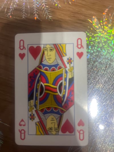 A close-up, high-angle, slightly blurry, eye-level shot shows a single playing card, the Queen of Hearts, placed on a wooden surface. The card is white with red and yellow detailing. A reflective, glittery surface with holographic elements is visible to the right and top of the card, creating bright glare. The card is oriented vertically, with the Queen's head facing upwards. The wooden surface appears to be a table with a visible grain pattern. The scene is indoors, with no visible windows or doors. The image contains no people. The location is Rosenovo, Bulgaria.