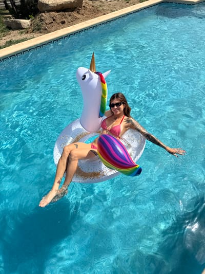 A woman wearing a red and white polka dot bikini and sunglasses floats on an inflatable unicorn in a swimming pool. The pool water is a vibrant blue, with sunlight creating patterns on the surface. The woman is positioned on the unicorn's body, with her legs extended and partially submerged in the water. She has tattoos on her left arm and leg. The unicorn float is white with gold glitter accents, a rainbow-colored mane, and a golden horn. In the background, the edge of the swimming pool is visible, constructed with light-colored coping stones and patterned tiles. Beyond the pool, there is a dry, earthy area with some rocks and sparse vegetation, suggesting an outdoor setting in a dry climate. This scene is located at a private residence with a swimming pool in Sant Vicenç de Montalt, Spain.