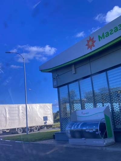 The image depicts a gas station with a "Magaza" sign and a logo featuring a red flower. In the foreground, a display unit containing car tires is visible. To the left, a large white truck trailer is parked on the side of the road. A street lamp stands prominently in the mid-ground. The background shows trees and a bright blue sky with some clouds. The scene is likely located in a suburban or rural area, given the presence of the truck and open space. The specific location in Заглядовка, Russia, is inferred from external information, as there are no explicit visual cues in the image to confirm this.