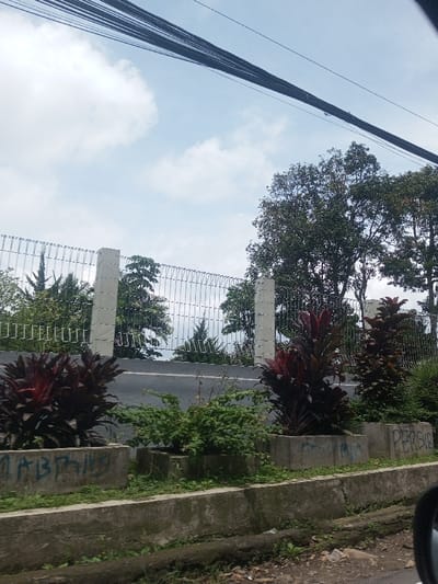 This image depicts an outdoor street scene in the city of Chori, Nigeria, under a partly cloudy sky. Numerous dark electrical or communication cables span horizontally across the top of the frame. Below these, a light-colored metal mesh fence, supported by two visible rectangular concrete posts, runs along the midground. Various green trees and foliage are visible both behind the fence and to its right. A dark asphalt road traverses the scene horizontally at mid-level. In the immediate foreground, a series of rectangular concrete planters are aligned along the road's edge. These planters contain a mix of green shrubs and plants with distinct dark red-purple foliage. Graffiti is present on some planters; the text "MABTANG" is partially visible on a planter to the left, and "PERSIB" is visible on a planter to the right. A concrete curb and a narrow drainage channel separate the planters from the rough dirt ground in the bottom foreground, which contains minor debris. A partial view of a vehicle's side mirror is discernible in the bottom right corner, suggesting the photograph was taken from within a vehicle. No individuals or active interactions are visible in the scene.