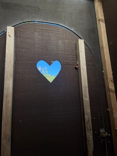 The image presents an interior perspective from a structure located in Sint-Pieters-Leeuw, Belgium. A large, dark brown panel, exhibiting a subtly textured surface, occupies the central view. Centered within this panel is a precisely cut, heart-shaped aperture, offering a view of a bright blue sky and sunlit green and yellow-green foliage beyond. The panel is vertically framed on both the left and right by weathered, unpainted wooden support beams, showing visible grain and minor imperfections. Above the main panel, a dark, arched section of the structure is visible, with a faint blue line tracing its upper edge. On the right side, adjacent to the lower portion of the right wooden beam, a metal door latch mechanism is partially discernible. A small reddish-brown object is lodged near the right beam, just above the heart cutout. Fine strands consistent with cobwebs are present on the extreme right edge of the scene. The interior space appears dimly lit, contrasting sharply with the brightly illuminated exterior observed through the cutout. No individuals or specific actions are depicted.