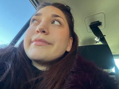 An image taken inside a vehicle in Dorlisheim, France, depicts a young adult female from the chest up, positioned towards the front passenger side. She possesses light-toned skin, dark brown hair with auburn highlights, and wears visible mascara on long eyelashes and a clear lip gloss. Her gaze is directed upwards and to the right of the frame, with a slight, closed-lip smile. She is dressed in a dark garment beneath a dark red, possibly faux fur, outer layer. The vehicle's interior features a light-colored headliner, a visible seatbelt retraction mechanism, and a black seatbelt strap extending downwards to the right. A dark seatback is partially visible. Bright blue sky is present through a window to the upper left, indicating daylight conditions.