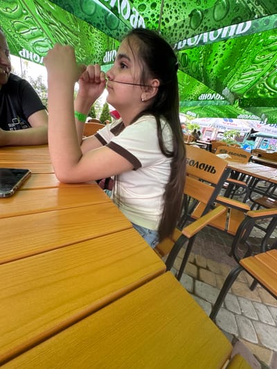 In Zaliznychne, Ukraine, under a green, droplet-patterned umbrella at an outdoor seating area, a young girl with long, dark hair is focused on manipulating a thin black string between her fingers and nose. She is wearing a white shirt with brown trim and a green wristband. To her left, another person is partially visible, wearing a black t-shirt. The scene is set at a wooden table, with a smartphone resting on it. In the background, several wooden chairs with black metal frames are arranged around tables, and the ground is paved with cobblestones.