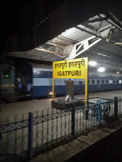 The image shows a train at Igatpuri railway station in Igatpuri, India. A yellow sign with the text "Igatpuri" in both English and Devanagari script is prominently displayed. A blue and white train is stopped at the platform. The platform is bordered by a metal fence. The station has a covered roof structure. The scene is dark, suggesting it is nighttime.