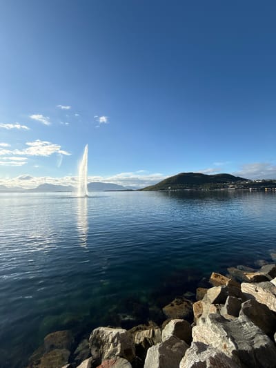 This is a wide shot of a body of water with a tall fountain in the center, located in Harstad, Norway. The water is a deep blue, with some lighter shades reflecting the sky and clouds. The fountain shoots a powerful stream of water high into the air, creating a white plume that catches the sunlight. In the background, there is a green, tree-covered hill with buildings clustered on its slopes and along the shoreline. Mountains are visible in the far distance, with a clear blue sky above, dotted with a few white clouds. In the foreground, large rocks and boulders line the edge of the water.