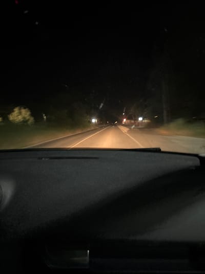 The image, rotated 90 degrees counter-clockwise for proper viewing, captures a nighttime scene from inside a vehicle, likely a car, in Parvomai, Bulgaria.
**Subjects:** The foreground, occupying the right side of the image, is dominated by a dark, textured surface, which appears to be the interior panel of a car door or dashboard. A small, metallic, or reflective object, possibly a handle or trim, is faintly visible towards the far right edge. Stretching horizontally across the mid-ground is an illuminated strip, clearly a road or path, lit by vehicle headlights or distant streetlights, casting a warm, yellowish glow. To the left of this road, a dark, indistinct area suggests an unlit environment or night sky, with some faint, dark green patches closer to the road, indicating roadside vegetation. In the distance along the illuminated path, several small, blurred light sources are visible, possibly from other vehicles or distant structures.
**Activity or event taking place:** The scene depicts a view from a moving or stationary vehicle at night, looking out onto a road.
**Notable details:** The time of day is clearly night, characterized by the deep darkness and the reliance on artificial lighting. The artificial illumination on the road creates a strong contrast with the dark surroundings. The overall image quality is dark and slightly indistinct, typical of low-light photography. No people are visible.
**Visible text:** There is no visible text in the image.