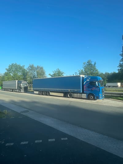 The image shows a roadside view in Ganderkesee, Germany. Two semi-trailer trucks are parked or stopped on a paved area next to a road. The truck on the left is dark gray and has "TRANSMAREK" written on its side. The truck on the right is bright blue with a graphic design on the cab and a blue trailer. The sky is clear and blue with a faint crescent moon visible. Trees and greenery line the background. The foreground shows a paved area with markings.