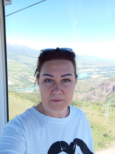 A woman is taking a selfie inside a cable car. She has dark hair, blue eyes, and sunglasses perched on her head. She's wearing a white t-shirt. Outside the cable car window, a mountainous landscape is visible, with greenery, rock formations, and a river winding through the valley. The location is Chinarkent, Uzbekistan.