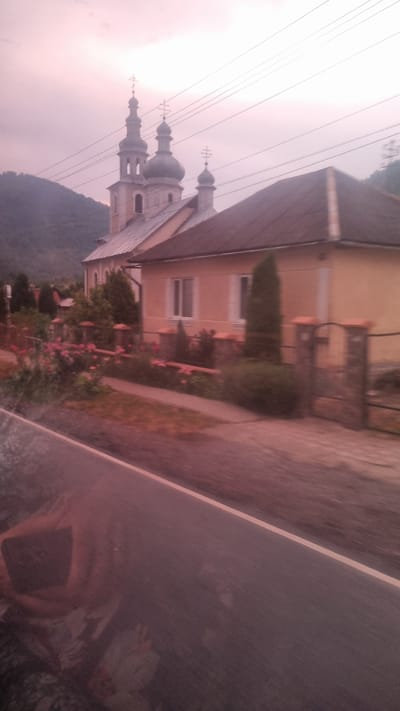 This is a photograph of a church with multiple domes and crosses, likely in Sil, Ukraine. The church is a prominent subject, with its towers reaching towards a hazy, pale sky. A building adjacent to the church, possibly a residential house or another church-related structure, is visible with light-colored walls and dark window frames. The scene is framed by greenery, including trees and a flowerbed with pink blossoms, suggesting a natural, possibly rural or suburban, setting. A paved pathway runs alongside the building and the garden. Power lines are visible crossing the sky, indicating the presence of infrastructure. In the foreground, a glossy, reflective surface, possibly a window or a piece of glass, obscures a portion of the scene and shows a distorted reflection of what appears to be a person holding a device and possibly other figures. The lighting suggests it is either early morning or late evening, with the sky being a soft, muted pink. The overall mood of the image is serene, with a touch of mystery due to the obscured foreground. No visible text is present in the image.