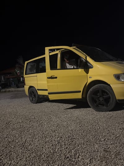 The image depicts a yellow Mercedes-Benz Vito van parked on a gravel surface at night. The driver's side front door of the van is open, revealing a young man sitting in the driver's seat, looking down. The van is facing left, with its rear end slightly angled towards the viewer. In the background, a building with a red roof is visible, along with some trees and a street light. The scene appears to be in a rural or suburban area. The overall lighting suggests it is nighttime, with artificial lights illuminating the van and its surroundings. The exact location in Zlataritsa, Bulgaria, is not specified beyond the context provided.