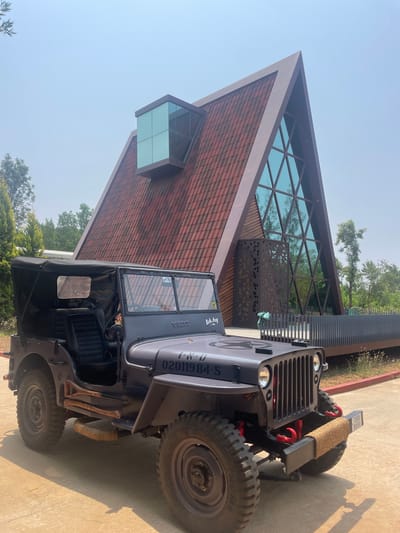A grey, vintage-style Jeep, possibly a customized Willys, is parked on a paved surface next to a modern building under a clear, bright sky. The Jeep features a classic slotted grille, round headlights, and a front bumper equipped with red tow hooks and a rope-wrapped object, possibly part of a winch. "WILLYS" is stenciled on the hood, and a "Bad Boy" sticker is visible on the windshield frame. A vertical registration number "02011984-S" is painted on the side of the engine compartment, and a license plate "CVN 0957" is on the front bumper. The vehicle has rugged off-road tires, a black soft top that appears to be rolled back, and black seats. Tools resembling a shovel and an axe are strapped to the side near the rear wheel.
The building behind the Jeep showcases contemporary architecture with a prominent triangular or pyramidal roofline. Its lower facade is covered in reddish-brown tiles or patterned wooden shingles. The upper section features a large glass facade with a dark geometric grid, reflecting the sky and surrounding trees. A distinctive, small, cube-shaped glass protrusion extends from one side of the building. A dark metal screen with geometric cutouts runs along a portion of the structure. The setting is outdoors, brightly lit by sunlight, casting strong shadows on the ground. Trees and green foliage are visible around the building. No specific activity is depicted; the scene is static.