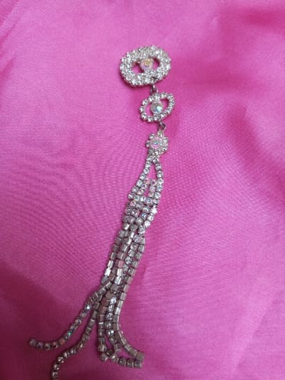 The image displays a silver-toned, rhinestone-encrusted piece of jewelry, possibly a brooch or decorative embellishment, laid flat on a vibrant pink fabric. The jewelry features a primary oval motif at the top, followed by two smaller oval and circular motifs, all adorned with numerous clear and iridescent rhinestones. From the lowest motif, multiple strands of rhinestones extend downwards, forming a tassel or fringe-like design. Some strands consist of tightly packed square settings, while others are single lines of rhinestones. The pink background fabric exhibits soft folds and a subtle sheen, suggesting a smooth material like satin or silk. The scene is located in Qarabucaq qəsəbə, Azerbaijan. No people, specific actions, or interactions are present.