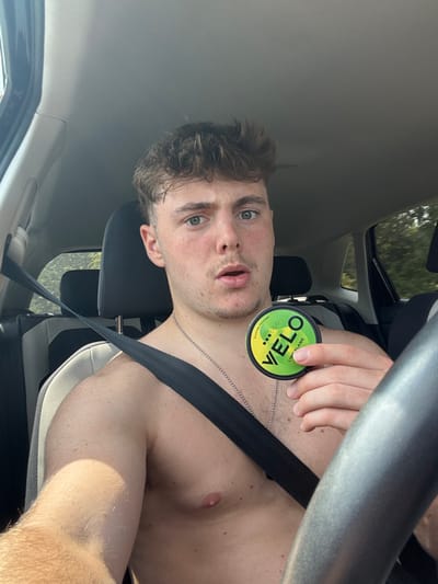A shirtless young adult male is seated in the driver's seat of a vehicle, described as being in Tickton, United Kingdom. He possesses light skin, short brown wavy hair, and light eyes, and looks directly at the camera with a slightly open mouth and a curious expression. A black seatbelt is properly fastened across his chest. He wears a thin silver chain necklace. In his left hand, he holds up a circular, green and black container marked "VELO LIME FLAME" and "Nicotine Pouches." The vehicle's interior features light and dark grey upholstered seats and a partial view of a dark grey steering wheel. Through the side window, blurred green foliage is visible, indicating an outdoor environment during daylight.