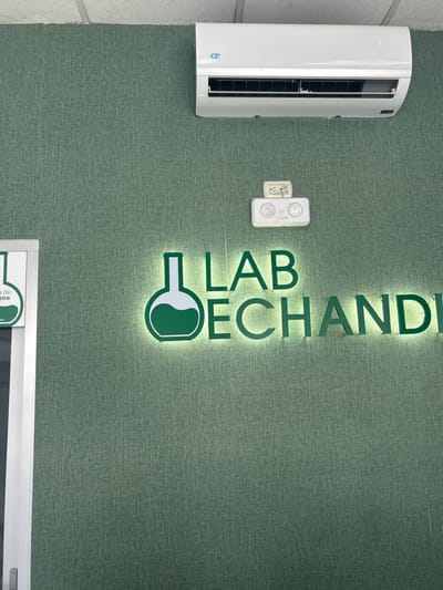 The image shows a wall with a green textured finish in a building located at the coordinates 9.94 latitude and -84.156 longitude in San Rafael, Costa Rica.  A white air conditioner unit is mounted on the wall. Above the air conditioner, a white rectangular box with a light fixture is mounted. A sign with the words "LAB ECHANDI" in green letters, illuminated from behind, is mounted on the wall.  The sign depicts a beaker next to the word "LAB". To the left of the sign is a white door frame with a sign that says "a de".  There are no people visible in the image.