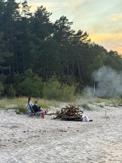 This is a vertically oriented, full-shot, outdoor photograph taken at dusk. The scene depicts a beach with fine, light-colored sand, bordered by a dense forest of pine trees and some dry grass. In the foreground, two people are seated in camping chairs on the sand, facing away from the camera and towards a bonfire. The bonfire is made of dry branches and is actively burning, producing a plume of white smoke that drifts upwards and blends with the twilight sky. One person is wearing a blue and white striped shirt and dark shorts, while the other is wearing a dark jacket and a blue and white striped shirt. A white plastic bag with red trim is placed on top of some driftwood near the bonfire. The sky is a pale yellow and orange, indicative of a sunset. The overall atmosphere is peaceful and relaxed, suggesting a quiet evening by the fire on the beach. The location is identified as Plieņciems, Latvia. There is no visible text in the image.