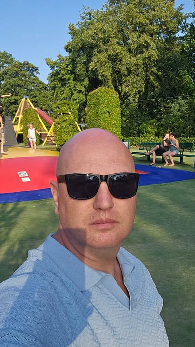 A bald, fair-skinned man wearing sunglasses and a light blue collared shirt is taking a selfie outdoors. In the background, a playground structure with yellow and red components is visible, with a young girl standing near it. Further back, two people are sitting on a green bench. The scene appears to be a park or recreational area in the city of Groenlo, Netherlands, given the presence of a playground and lush greenery. The lighting suggests it is daytime, likely late afternoon or early evening due to the long shadows.