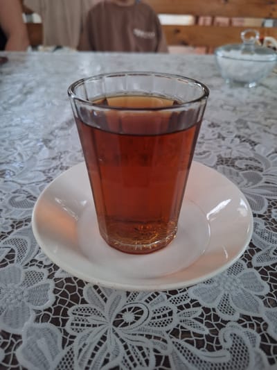 A medium shot shows a glass of iced tea on a white saucer, which rests on a white lace tablecloth patterned with a dark brown background. The glass is about three-quarters full of dark amber liquid with ice cubes visible near the bottom. The glass has a faceted exterior. The saucer is round with a slightly raised rim. Behind the glass, a clear glass sugar bowl filled with white sugar is visible. In the background, two blurry figures are seated at a wooden table. The scene appears to be in a cafe in Nabran, Azerbaijan.