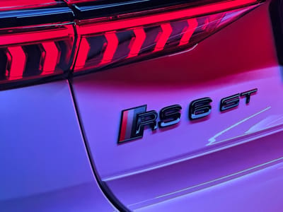 The image is a close-up shot of the rear of an Audi RS6 GT. The taillights are illuminated with a distinctive chevron pattern in red. The "RS6 GT" badge is prominently displayed on the car's body, featuring a black finish with a stylized Audi Sport stripe on the "RS" portion. The car's bodywork reflects ambient lighting, which appears to be a mix of red and purple hues, suggesting it might be displayed in a showroom or under colored lighting. The setting is not identifiable as Bezden, Bulgaria, or any specific city. There are no people or actions depicted.