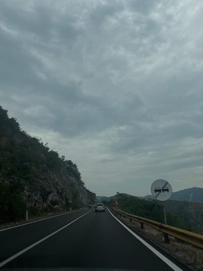 The image shows a road with a mountain on the left and a mountain range on the right. There are a few cars on the road, and a traffic sign that indicates no overtaking. The sky is cloudy and overcast. The road appears to be a two-lane highway. The location is identified as Milojevići, Montenegro. The scene is likely captured from inside a moving vehicle.
