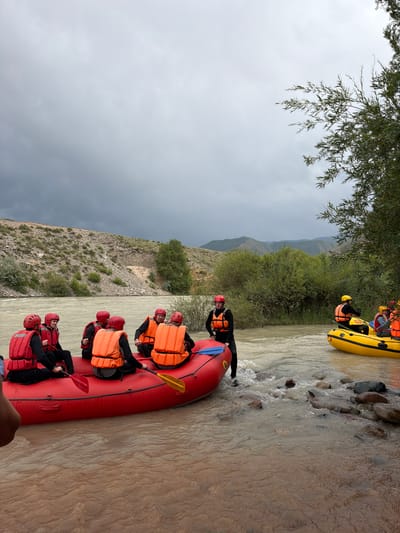In Sasyk-Bulak, Kyrgyzstan, a group of people are preparing for a rafting excursion on a cloudy day. Two inflatable rafts are in the water near the shore. A large red raft, with six individuals wearing red and orange life vests and helmets, is partially submerged in the shallow water. Some individuals are holding paddles. To the right, a smaller yellow raft, with two individuals also wearing life vests and helmets, is further out in the river. The river water is murky brown, with visible rocks in the foreground. The surrounding environment consists of a rocky, vegetated hillside and lush green trees and bushes. In the distance, rolling hills under a gray, overcast sky complete the scene.