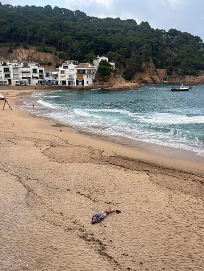 The image shows a sandy beach in Tamariu, Spain, located at the coordinates 41.918 latitude and 3.207 longitude. The beach is mostly empty except for a man laying face down on the sand in the lower right portion of the image. His arms are extended to his sides, and he is wearing a short-sleeved, blue and white patterned shirt. There are a few footprints and tire tracks in the sand.  In the distance, there are white houses built into a rocky cliff covered in green trees. Behind the houses, a blue-green body of water stretches out, with a small, dark-colored boat visible in the distance. The water is relatively calm, with a few small whitecaps. The sky is overcast and gray.