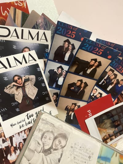 The image shows a collection of items scattered on a light-colored surface. Dominating the scene are several copies of "DALMA Magazine," each featuring a woman with sunglasses in a tan trench coat. The magazines advertise Yerevan Fashion Week. There are also photo booth-style prints labeled "Class of 2025," depicting groups of young people in graduation attire. A red card with "PR" visible is present, alongside a Polaroid-style photograph of a person in a striped shirt. A framed photograph of two people and a card with a quote "YOU ARE CAPABLE OF AMAZING THINGS BECAUSE YOU ARE AMAZING" are also visible. There are no indications suggesting that it is related to the city of Aratashen, Armenia.