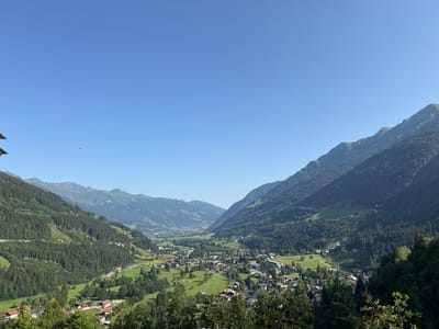 The image shows a panoramic view of the town of Bad Gastein, Austria, nestled in a valley surrounded by lush green mountains. The sky is a clear, vibrant blue, with a few small birds visible in the upper left quadrant. The mountains are densely covered with dark green pine trees, with some areas showing patches of lighter green grass and exposed rock. The town itself is a collection of buildings with red and brown roofs, scattered across the valley floor and on the lower slopes of the mountains. Some of the buildings appear to be residential homes, while others suggest commercial or hotel establishments, characteristic of a resort town. A golf course with manicured green fairways is also visible in the middle ground, adding to the sense of a developed recreational area. The overall impression is one of a peaceful, scenic mountainous landscape with a charming European town.