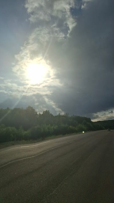 A bright, low sun shines through dramatic, dark clouds, casting sunbeams onto a verdant treeline and a two-lane asphalt road. The road curves to the right, with a white line marking the edge of the lane. A single white car is visible in the distance on the right side of the road. The environment appears to be rural, possibly on the outskirts of Budukan, Russia. The time of day is likely late afternoon or early evening, given the low angle of the sun and the interplay of light and shadow. The overall mood is one of a transitional weather event, with the sun breaking through an impending storm.