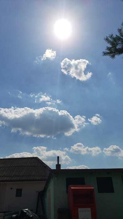 This media file captures a bright, sunny day with a clear blue sky, dotted with fluffy white clouds. The sun is prominently visible, casting a strong glare and creating lens flare effects. In the foreground, the upper branches of a coniferous tree are visible against the sky.
On the right side of the image, a building with a light green facade is partially seen. Attached to this building is a red, rectangular object, possibly a mailbox or a receptacle of some sort, with some visible text on its side. The text appears to be in a European language and reads "Pošta a zberné miesta" and below that "Obec Vyšné Nemecké, Hrusov." This suggests the location is in Vyšné Nemecké, Slovakia, and the object is related to postal services or collection points in the village.
Beneath the green building, another structure with a corrugated metal roof is visible. This building appears to be a house or another type of dwelling, with a small, dark-framed window visible on its lighter-colored wall. The overall scene is one of a rural or semi-urban setting on a pleasant, sunny day. The time of day is clearly daytime, given the intensity of the sun and the brightness of the sky. No people are visible in the frame. The mood evoked is peaceful and ordinary, depicting a typical day in the described location.