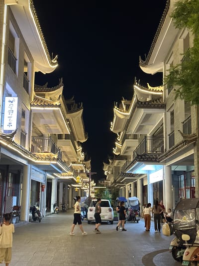 In the city of Eshan Yi Autonomous County, China, at night, a pedestrian street is illuminated by decorative lights along the eaves of traditional Chinese-style buildings. The buildings feature multiple stories with balconies and ornate roofs. Several people are walking on the paved street, and a few scooters and a white car are parked along the side. A young child in a light-colored outfit stands facing away from the camera, looking towards the street. The atmosphere is lively and illuminated, suggesting it's a popular area for evening strolls.