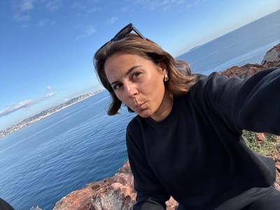 A female individual with medium brown hair, fair complexion, and dark eyes is depicted in a close-up selfie, posing with puckered lips directly towards the camera. She is wearing a black crew-neck sweatshirt, dark trousers, and gold-colored hoop earrings; black sunglasses are positioned atop her head. The subject is situated on reddish-brown rock formations, overlooking the deep blue Mediterranean Sea from a high vantage point in Théoule-sur-Mer, France. The background features a long, distant urbanized coastline extending along the horizon under a clear, bright blue sky with minor cloud formations. A small, indistinct landmass or structure is visible on the far right horizon.