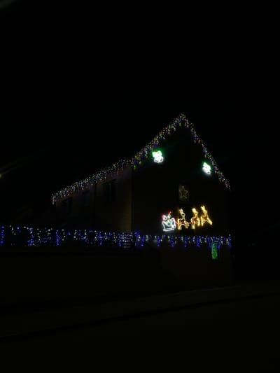 A building is adorned with Christmas lights against a dark night sky. Multi-colored lights outline the roof's edge. Illuminated figures, including reindeer, Santa Claus, and stars, decorate the building's front. A string of blue lights runs vertically along the building's side. Some windows are visible, adding depth to the structure. The overall effect is festive and celebratory.