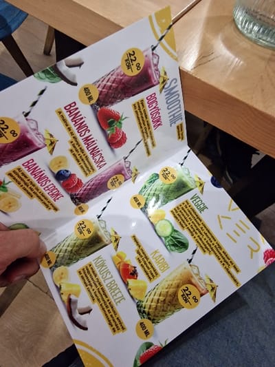 The scene depicts an indoor setting, likely a cafe or restaurant, in Odorheiu Secuiesc, Romania. A light-skinned hand with visible finger hair is holding open a folded, glossy, laminated menu on the lower left. The menu prominently features various smoothie options with their names in Hungarian, alongside colorful illustrations of the drinks and their ingredients, prices uniformly listed at "22 RON", and volumes specified as "400 ML". Visible smoothie titles include "BOGYÓSOKK" (Berries), "BANÁNOS MÁLNÁCSKA" (Banana Raspberry), "BANÁNOS EPERKE" (Banana Strawberry), "KOKUSZ BREEZE" (Coconut Breeze), "KARIBI" (Caribbean), and "VEGGIE". The ingredients depicted include strawberries, raspberries, blueberries, bananas, pineapple, coconut, papaya, mango, cucumber, and spinach. A light-colored wooden table with a visible grain occupies the right side of the frame. On this table, a clear, textured glass containing a transparent liquid is partially visible at the top right. Dark blue fabric, consistent with denim, is visible in the lower section of the image, presumably belonging to the person holding the menu.