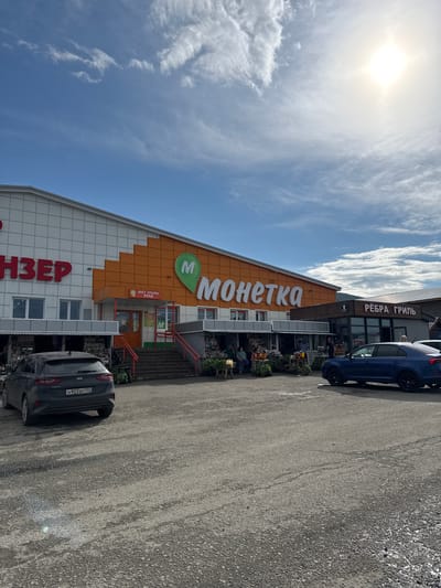 The image depicts a storefront in Inzer, Russia, featuring a "Monetka" supermarket and a "Rebra Gril" (Ribs Grill) establishment. The facade of the Monetka store is prominent, with its orange and white design and a large green logo with a white "M". The entrance to Monetka has stairs leading up to it, and a sign indicates "ВХОД" (Entrance). Next to Monetka, the Ribs Grill has a darker wooden exterior with red lettering. In the foreground, a gray Kia Ceed station wagon is parked, facing away from the camera, displaying a license plate ending in "702". The ground is a paved surface, appearing to be asphalt or gravel, with some visible cracks and damp patches. Several people are visible near the entrance of Monetka, some appearing to be customers browsing stalls set up outside, selling produce and other goods. A blue sedan is parked to the right of the frame, partially obscuring the Ribs Grill establishment. The sky is blue with scattered white clouds, and the sun is bright, casting a glare in the upper right portion of the image.