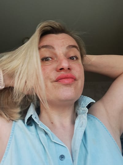 A close-up, low-angle portrait depicts an adult female positioned indoors. She has blond hair, parted slightly left, with strands falling across her forehead. Her face is light-complexioned, featuring visible freckles across the cheeks and nose, wide-open dark brown eyes with black eyeliner, and lips puckered into a 'kiss' shape, colored with pink lipstick. She wears a light blue, sleeveless, collared denim-style shirt, with the top button visible and unbuttoned. A light-colored smartwatch or fitness tracker is present on her left wrist. Both of her arms are bent at the elbow, with hands placed behind her head, partially obscured by her hair. The background consists of an indistinct dark grey ceiling or wall, with a small circular fixture partially visible in the upper left quadrant. No specific geographic features or landmarks indicative of the city of Алфёрово, Russia, are present within the frame.