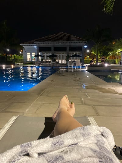 A poolside scene is captured at night, featuring a woman relaxing on a lounge chair near a pool. The pool's water reflects the lights of a nearby building, which has multiple stories and appears to be a resort or residential complex. Dark umbrellas are arranged around tables and chairs near the pool. The woman has her legs extended, with her feet sporting green nail polish. She is draped in a blue and white striped towel or robe. The background includes palm trees and other vegetation, enhancing the outdoor ambiance. The location is presumed to be near La Aurora, Ecuador.