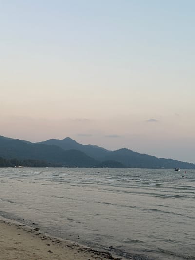 A scenic view in Baan Klong Prao, Thailand, shows the coastline meeting a silhouetted mountain range under a hazy sky. The sea is gently rippled, extending from the shore toward the distant mountains. A sandy beach borders the edge of the image. There is a boat located further out in the water.