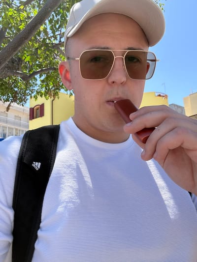 A man wearing a white hat, gold-framed sunglasses, and a white shirt with a black Adidas backpack strap is holding a brown vaping device to his lips. The photo appears to have been taken looking up at the man. Behind the man, a street scene is visible, including trees, a yellow building with red-shuttered windows, and a white building. This suggests the photo may have been taken in the city of Mola di Bari, Italy.
