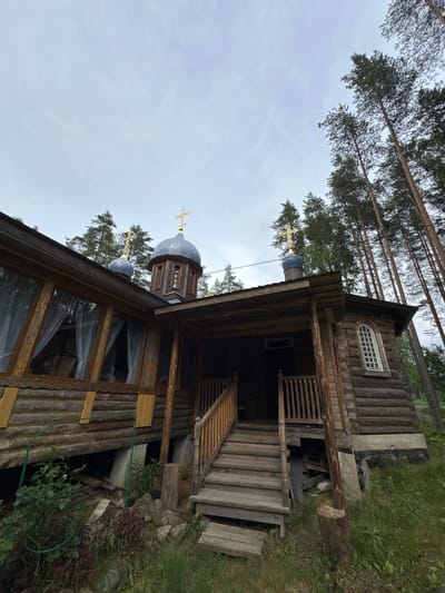 The image shows a wooden church in the village of Nenimäki, Russia. The church has two blue domes topped with golden crosses and a wooden facade. A wooden staircase leads to the entrance of the church. The church is surrounded by tall pine trees and lush green grass. The sky is overcast, and the overall atmosphere is peaceful and serene.
