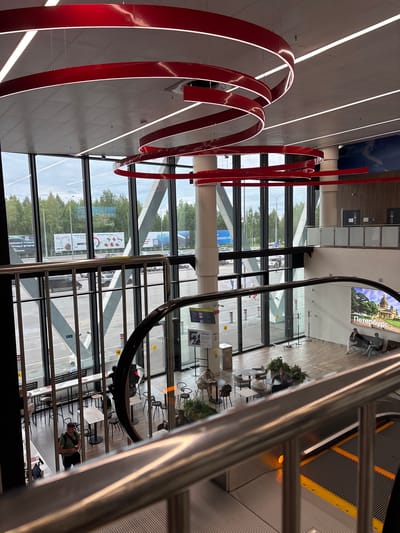 The image depicts the interior of a modern, multi-level building, likely an airport or train station, with large windows offering views of the outdoors. A prominent red, ribbon-like sculpture is a central decorative element. The scene shows various people interacting within the space. On the upper level, a large digital display shows an image of a domed building and the word "Петербург" (Petersburg). Several individuals are seated on a bench in front of this display.

On a lower level, accessed via an escalator, there is a seating area with tables and chairs, occupied by several people. Some are seated at tables, seemingly engaged in conversation or relaxing, while others are walking or standing. The overall atmosphere appears calm and functional, typical of a transit hub.

Through the large glass windows, an outdoor scene is visible, suggesting it is daytime with overcast skies. Trees and parked cars can be seen, indicating the building is situated in an urban or semi-urban environment. The architecture features clean lines, contemporary lighting, and polished surfaces. The time of day is not explicitly stated but appears to be daytime.

Visible text includes "Петербург" on the large display. Other text is present on signs and informational displays within the building and on advertisements visible through the windows, but it is mostly illegible or too small to decipher clearly in the given resolution. One sign visible through the window appears to be an advertisement with images and Russian text.