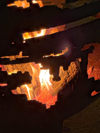 This is a close-up, medium shot of a bonfire within a fire pit, likely outdoors, possibly in the Слабадскі сельскі Савет region of Belarus. The image is dominated by the intense glow of the flames, which are a vibrant orange and yellow. The fire is consuming pieces of wood, some of which are charred and textured, showing intricate grain patterns. These wood pieces are partially obscured by a dark, silhouette-like metal structure that forms the rim of the fire pit. This metal structure has a rough, irregular edge, with cutouts that allow views into the burning fire. The background is mostly dark, with some hints of the surrounding environment, possibly a stone or gravel surface at the bottom right. The overall impression is one of warmth and controlled fire.