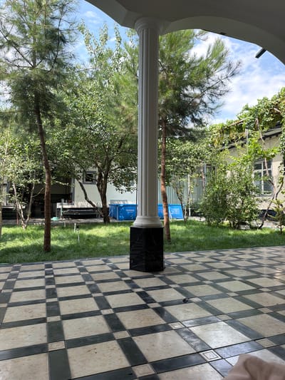 In Mindanabad, Uzbekistan, a courtyard scene unfolds. A white, fluted column stands prominently in the foreground, its base adorned with black marble. The ground is paved with a checkered pattern of light beige and dark grey tiles, with a light shadow cast on the tiles. Beyond the tiled area, a lush green lawn stretches out, dotted with various trees. A bright blue inflatable swimming pool, identified by the "Bestway Steel Pro" label, is visible in the mid-ground to the right of center. Behind the lawn and pool, a light-colored building with windows can be seen. The sky above is partly cloudy. The scene appears to be set in a residential or recreational outdoor space.