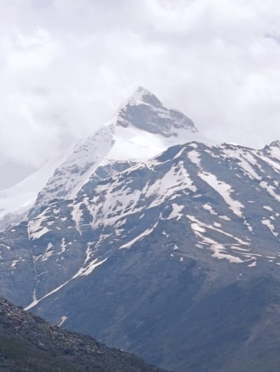 The image displays a majestic snow-capped mountain peak under a cloudy sky. The mountain's lower slopes are covered in dark, rocky terrain, transitioning to patches of snow and rock higher up, culminating in a pristine white summit. The clouds appear soft and diffused, creating a muted, atmospheric light. The scene evokes a sense of remote, natural beauty.