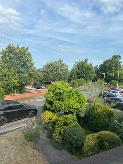 The image shows a residential street scene in West Lancashire, United Kingdom. The view is from an elevated position, possibly a window or balcony. The sky is partly cloudy. A row of parked cars lines the street, including a black SUV in the foreground and other vehicles of various colors further down the street. Lush greenery dominates the scene, with trees and bushes along both sides of the road. Streetlights are visible. The road surface is asphalt. A brick wall is also visible along one side of the street. There are no visible people in the scene.