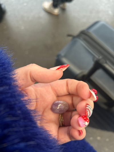 A human left hand, adorned with elaborate nail art and a gold-toned ring, is centered in the foreground, holding a smooth, tumbled purple or lavender-colored stone. The hand's thumb and index finger feature pointed, stiletto-shaped nails with clear bases, red tips, and silver glitter accents. The middle finger displays a clear nail with red swirls and silver lines. The ring finger has a clear nail with red heart designs and silver accents, and a gold-colored band with small facets or diamonds is worn on this finger. The pinky finger also has a clear nail with a red tip. The person is wearing a textured, royal blue faux-fur garment, visible on the left side of the frame.
The environment is an indoor public area, identified as being within the city of Bath, United Kingdom. The background is out of focus. To the upper right, a dark grey or black hard-sided suitcase with a visible handle is present. In the upper left background, the lower legs and feet of another individual are partially visible, wearing light-colored shoes. The floor surface appears to be a light grey, consistent with a public terminal or waiting area. No specific landmarks of Bath are discernible in the image.