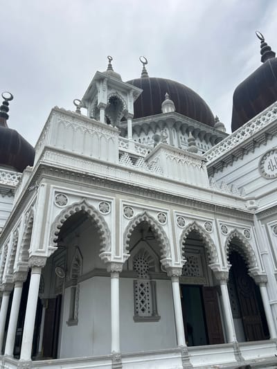 The image shows the Zahir Mosque in Alor Setar, Malaysia, viewed from a low angle looking upwards towards the sky. The mosque features a white facade with intricate architectural details, including arches, columns, and decorative patterns. Several dark domes, topped with crescent moons, are visible. The sky is overcast and grey. The architecture appears to be a blend of Islamic and Moorish influences. No people are visible in the frame. The mosque is a significant landmark in Alor Setar.