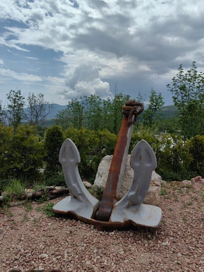 The image shows a large, gray and rust-colored anchor placed on a gravel-covered ground in Luchistoe, Ukraine. The anchor has two flukes and a stock with a looped chain at the top. Behind the anchor are trees, shrubs, and a rock, with a mountainous landscape and cloudy sky in the background.